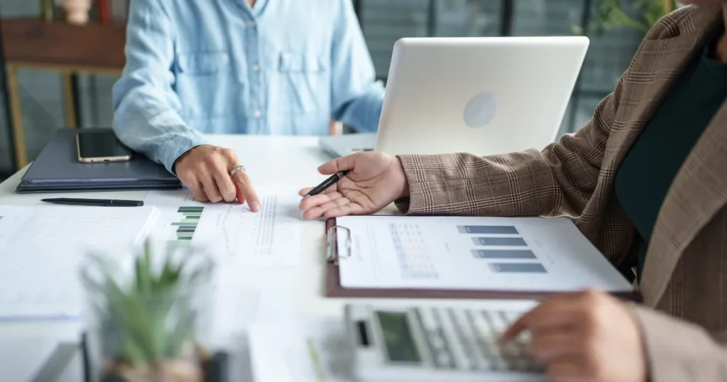 Two colleagues review business reports and charts together at a desk with a laptop and calculator, illustrating the Role of Internal Audit in financial oversight.